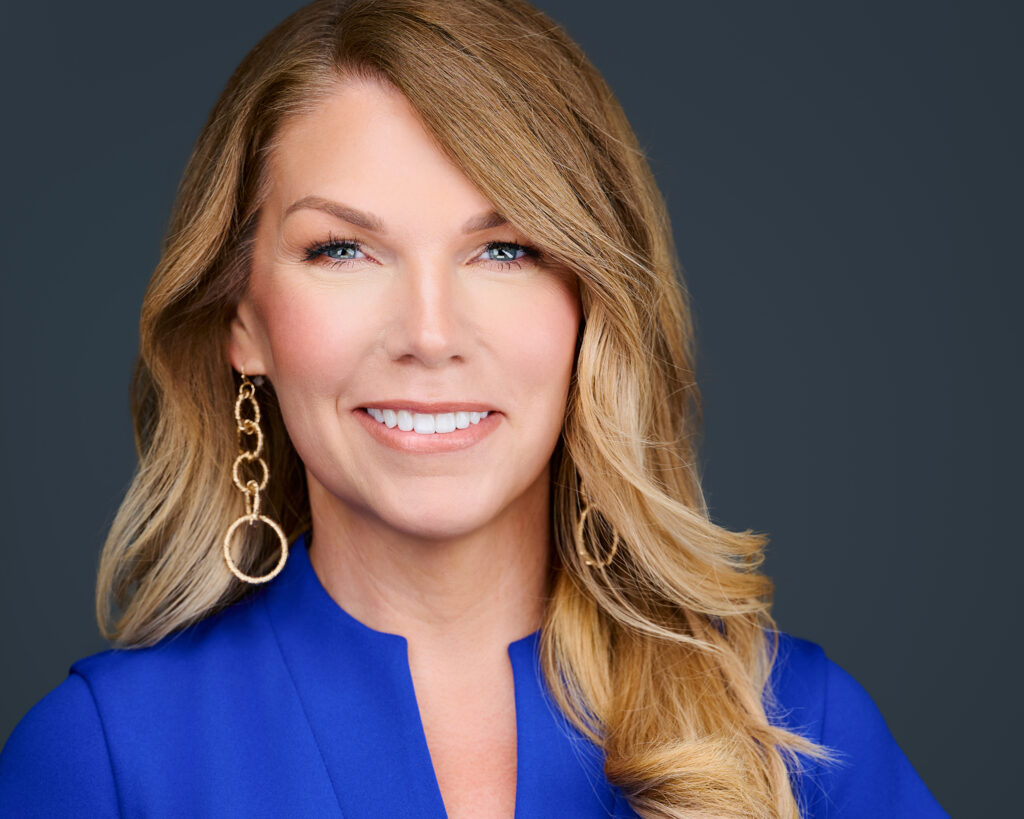 Professional headshot of a smiling REALTOR® wearing a royal blue blouse against a dark gray studio background in St. Louis, Missouri.