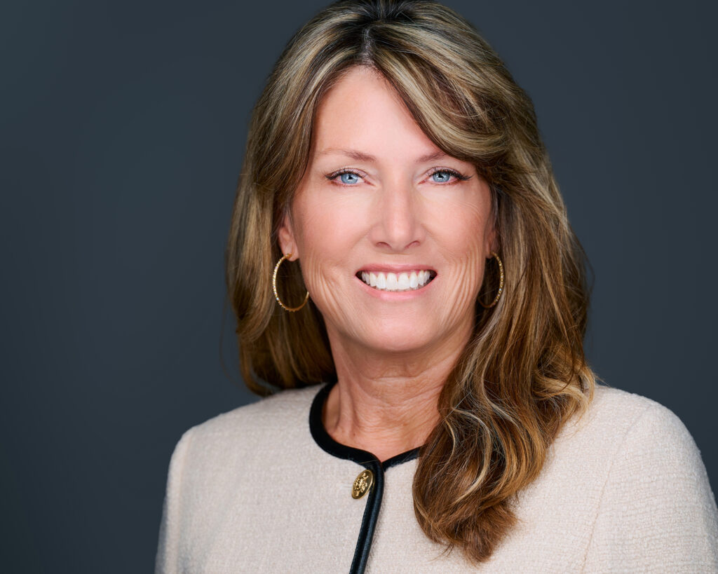 Professional headshot of a smiling REALTOR® wearing a cream blazer against a dark gray studio background in St. Louis, Missouri.
