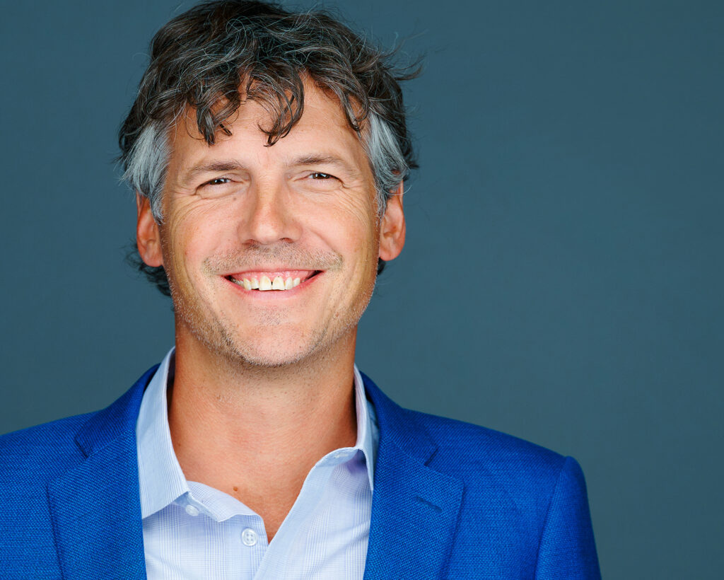 Professional headshot of a smiling man in a blue blazer against a blue-gray studio background