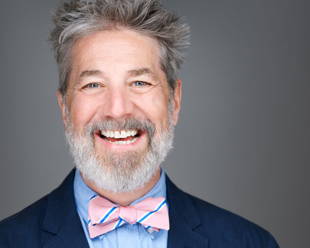 Professional executive headshot of a smiling man with salt-and-pepper hair and a full gray beard, wearing a blue suit jacket, light blue shirt, and pink and blue bow tie, photographed in a St. Peters, Missouri studio serving the St. Louis area.